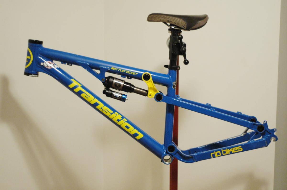 Transition Bottlerocket: A close-up image of a blue bicycle frame with yellow lettering, featuring the brand name "Transition" and the model name "Bottlerocket." The frame is showcased on a stand, highlighting the suspension system and seat post. The background is neutral to emphasize the bike frame.