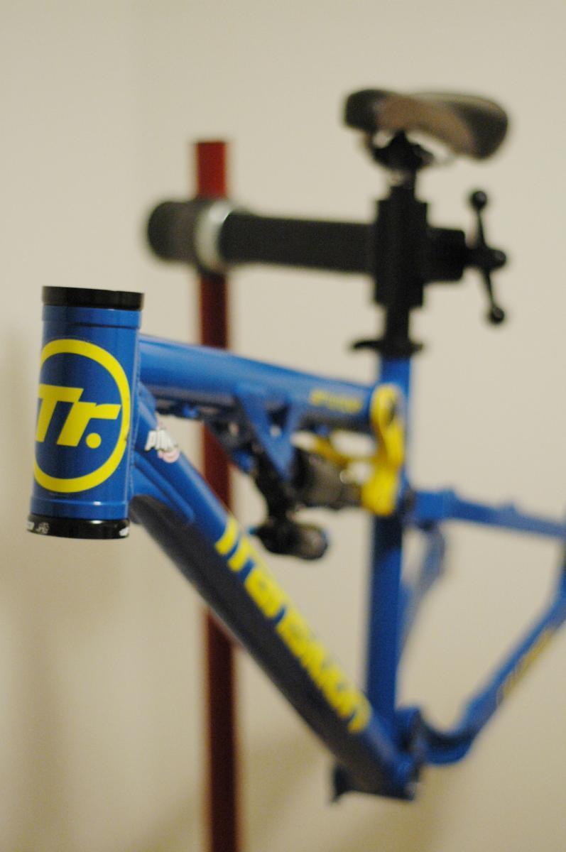 Transition Bottlerocket: Image of a blue bicycle frame with yellow accents, mounted on a repair stand. The focus is on the frame, showcasing its distinctive design and color. The background is blurred, emphasizing the bike frame.