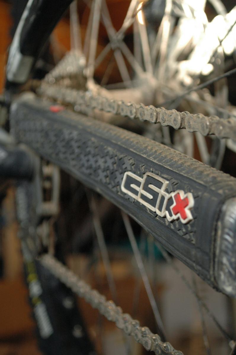 cSixx Chainstay Protector: Close-up of a bicycle chain and rear derailleur, showcasing dirt and wear on the components, with a focus on a branded chainstay protector. The background is slightly blurred, emphasizing the bike's mechanical parts.
