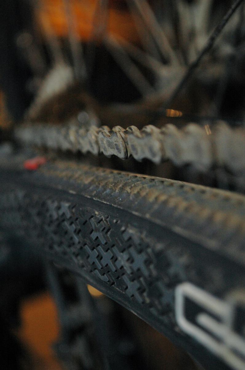 cSixx Chainstay Protector: Close-up view of a bicycle chain and tire, showing detailed texture and dirt accumulation, with a blurred background that hints at additional bike components.