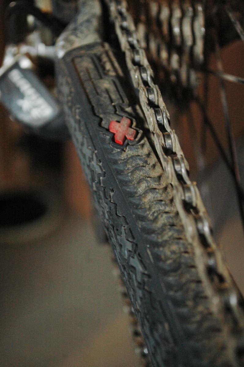 cSixx Chainstay Protector: Close-up of a mountain bike tire and chain, showing a dirty tread pattern and a small red cross symbol on the tire sidewall. The background is blurred, emphasizing the bike components.