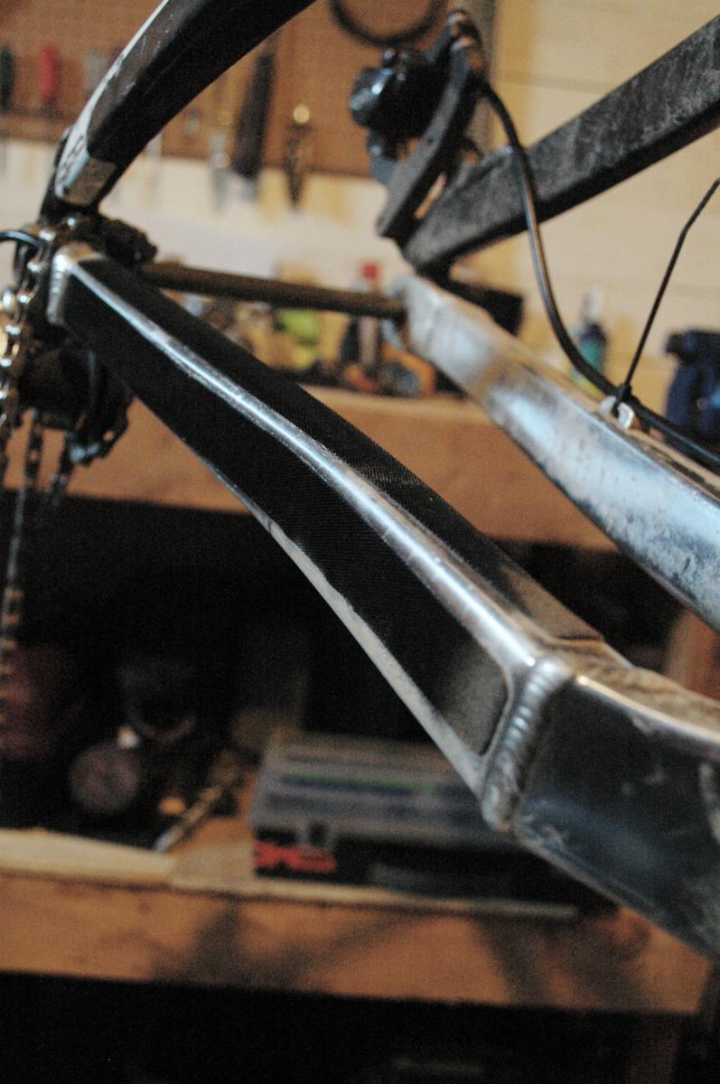 cSixx Chainstay Protector: Close-up view of a bike frame in a workshop, highlighting the welded metal structure and chain. The background features various bike tools and supplies on a workbench, suggesting a bike repair or assembly setting.