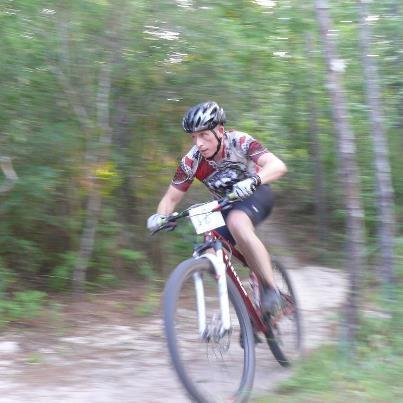 Niner EMD 9: A mountain biker in motion navigating a dirt trail surrounded by trees. The rider is wearing a helmet and a colorful cycling jersey, with a blurred background indicating high speed.