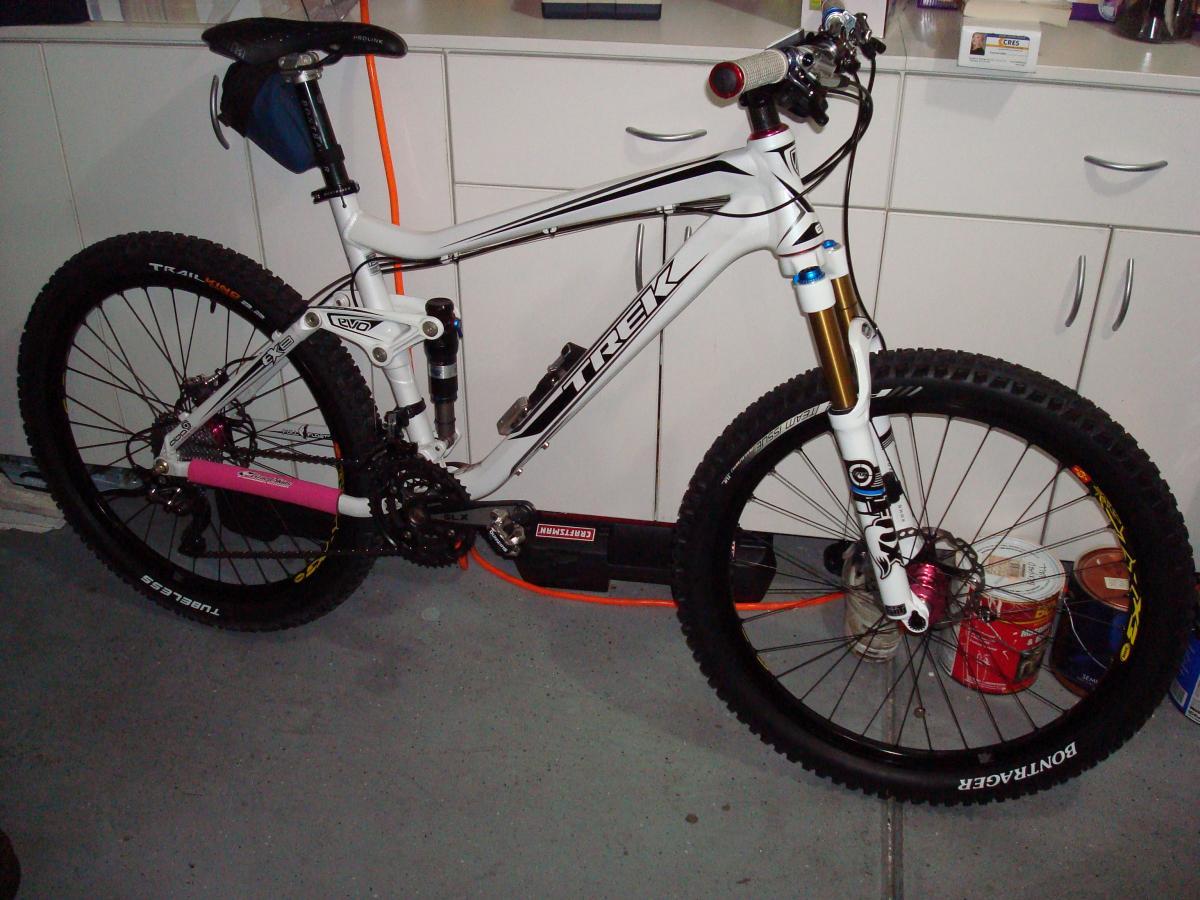 Trek Fuel EX 8: A white and black mountain bike is displayed in a garage setting. The bike features thick tires, a rear suspension system, and a pink chainstay protector. It is leaning against a white cabinet, with various tools and cans visible in the background. The bike is equipped with a distinctive fork and handlebars, showcasing its design for off-road cycling.