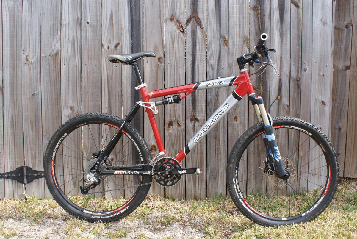 Michelin Wild Race'R: A red and black mountain bike is leaning against a wooden fence. The bike features thick tires, a front suspension fork, and a multi-speed crankset. The frame has "Rocky Mountain Slayer" written on it, with components showcasing a sporty design. Grass and dirt are visible at the base of the bike.