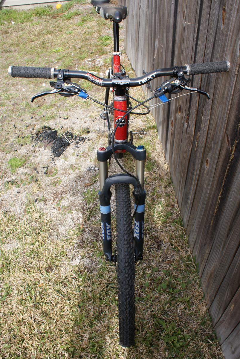 Michelin Wild Race'R: Alt text: A mountain bike viewed from the front, featuring black handlebars with textured grips, blue and red brake cables, and a red and silver frame. The bike has suspension forks and rugged tires, set against a grassy background with a wooden fence nearby.