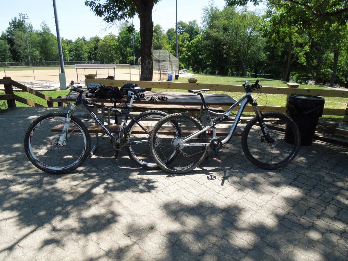 Gary Fisher Cobia: Two mountain bikes parked next to a picnic table in a sunny outdoor park setting, with a baseball field and trees in the background. A pair of gloves is resting on the table, and a trash can is visible nearby.