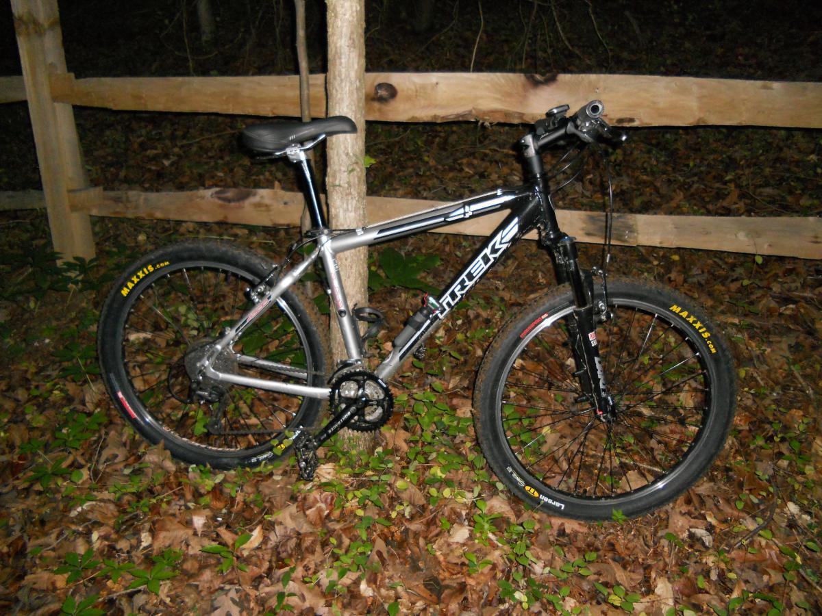 Maxxis Larsen TT UST: A black and silver mountain bike parked next to a tree in a wooded area, with fallen leaves and greenery visible on the ground. A wooden fence is in the background.