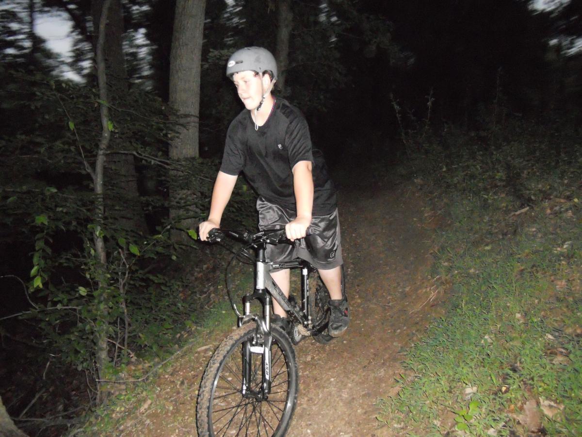 Trek 4300: A young person riding a mountain bike on a narrow dirt path in a wooded area during twilight. The cyclist is wearing a helmet and sports attire, and the surrounding environment is lush with greenery and trees.