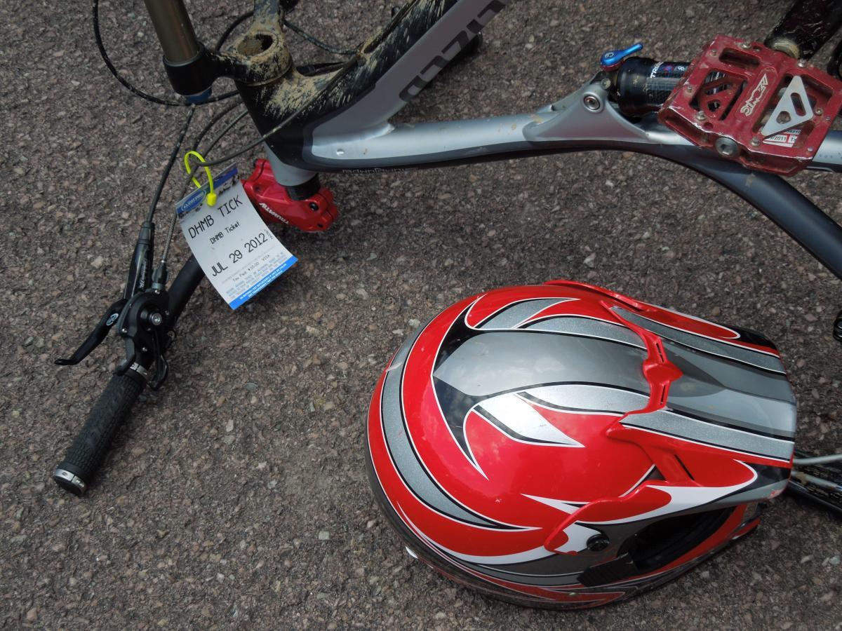 Specialized Stumpjumper FSR Comp 29: A close-up view of a mountain bike with a red and silver helmet nearby. The bike, partially covered in dirt, features a red brake lever and a tag labeled "DHMB TICK" dated July 29, 2012, indicating it might have been used for an event. The ground surface is gravel.