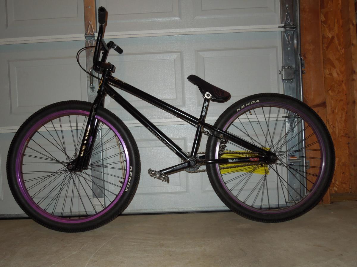 Kenda Small Block Eight: A black BMX bike with purple rims, leaning against a garage wall. The bike features a simple frame design, straight handlebars, and knobby tires suitable for riding on various terrains. The background shows a closed garage door.