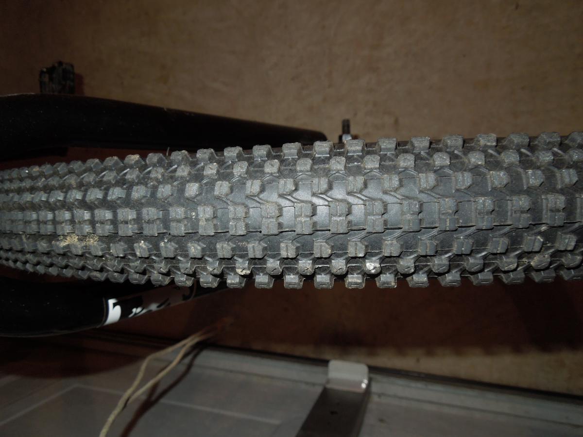 Kenda Small Block Eight: Cycling tire viewed from the side, showcasing a textured tread pattern designed for traction. The tire appears slightly worn with dirt particles visible on the surface, indicating outdoor use. The background is blurred, focusing on the tire and its details.