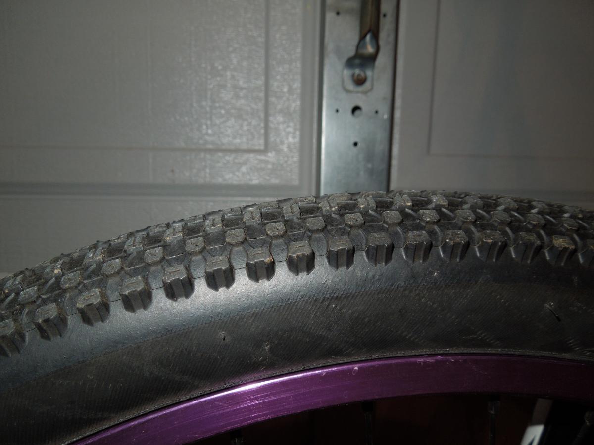 Kenda Small Block Eight: Close-up image of a textured bicycle tire, showcasing the tread pattern, with a purple rim visible at the bottom and a garage door in the background.