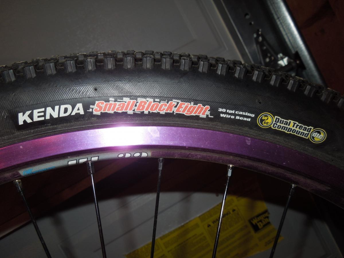 Kenda Small Block Eight: Close-up image of a Kenda Small Block Eight mountain bike tire, featuring a black rubber surface with visible tread patterns. The tire is mounted on a purple rim, and labels on the tire highlight specifications such as "30 tpi casing," "wire bead," and "Dual Tread Compound."