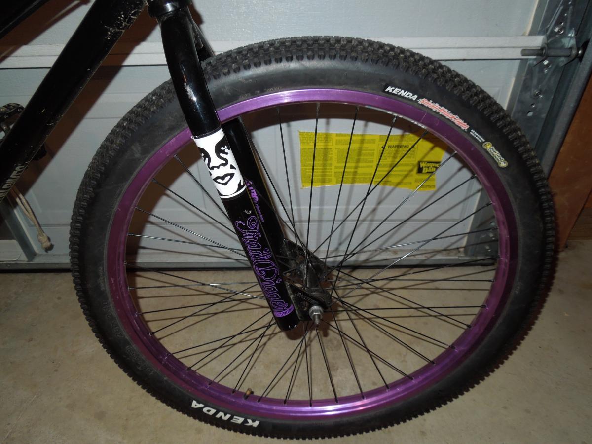 Kenda Small Block Eight: A close-up view of the front wheel and fork of a black bicycle, featuring a purple rim and Kenda tires. The fork displays a distinctive graphic design, while the background shows a garage setting with a warning label.