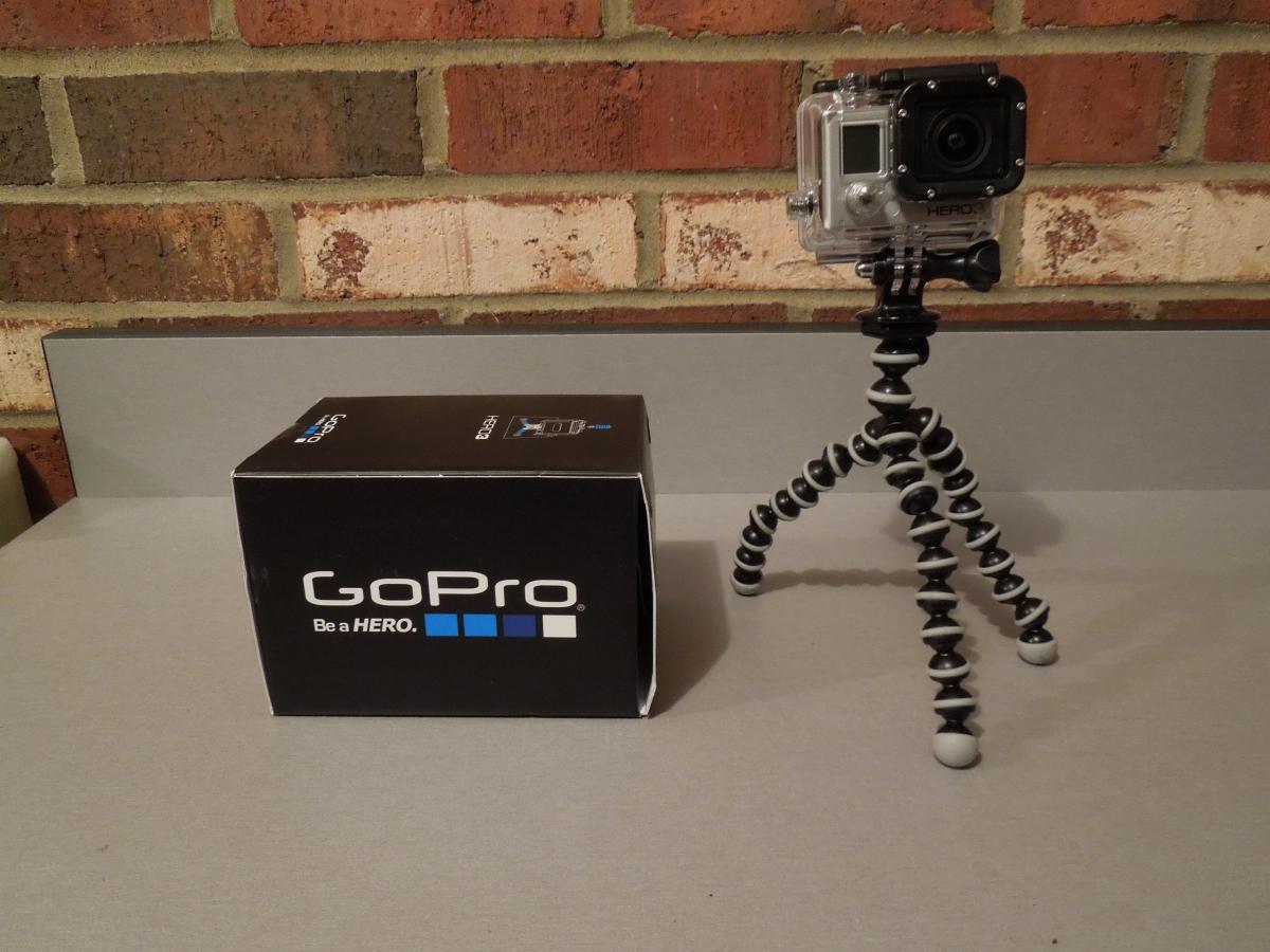 GoPro Hero3 Silver Edition: A GoPro camera mounted on a flexible tripod, positioned next to its original box, set against a brick wall background.