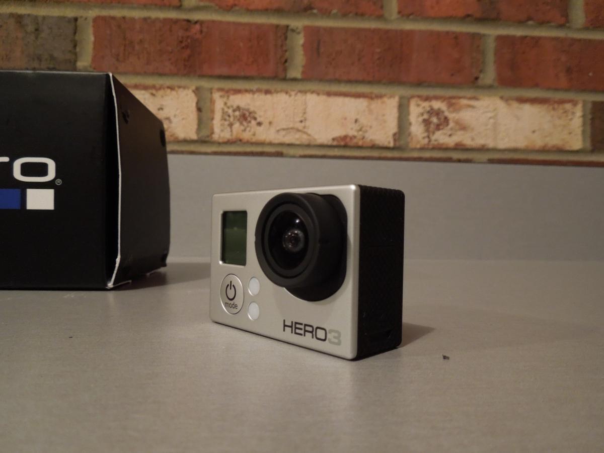GoPro Hero3 Silver Edition: A GoPro HERO3 camera positioned on a gray surface, with its lens and controls visible. A black GoPro box is partially in the background, with a brick wall behind it.