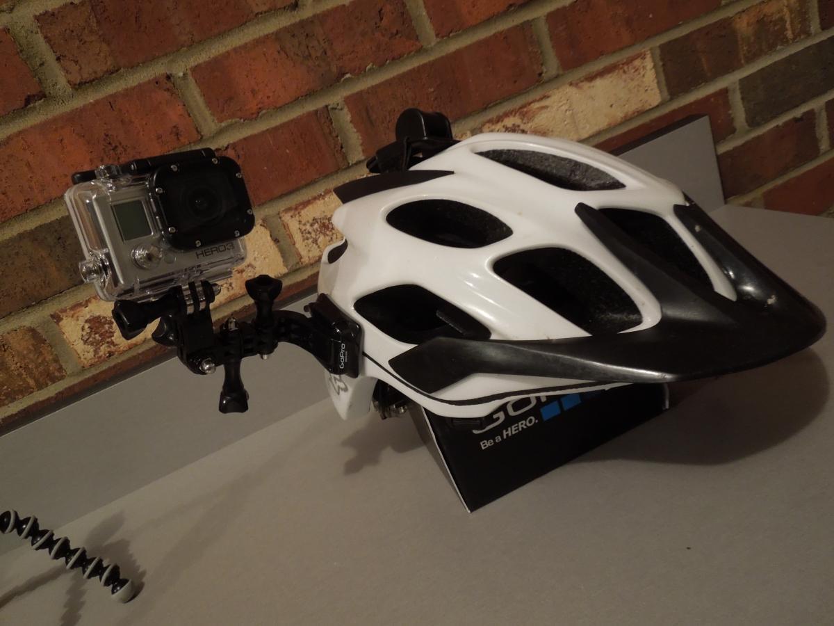 GoPro Hero3 Silver Edition: A white and black bicycle helmet mounted with a GoPro Hero3 camera, positioned on a gray surface against a brick wall backdrop. The helmet features ventilation holes and a sleek design, while the camera is attached securely, ready for recording.