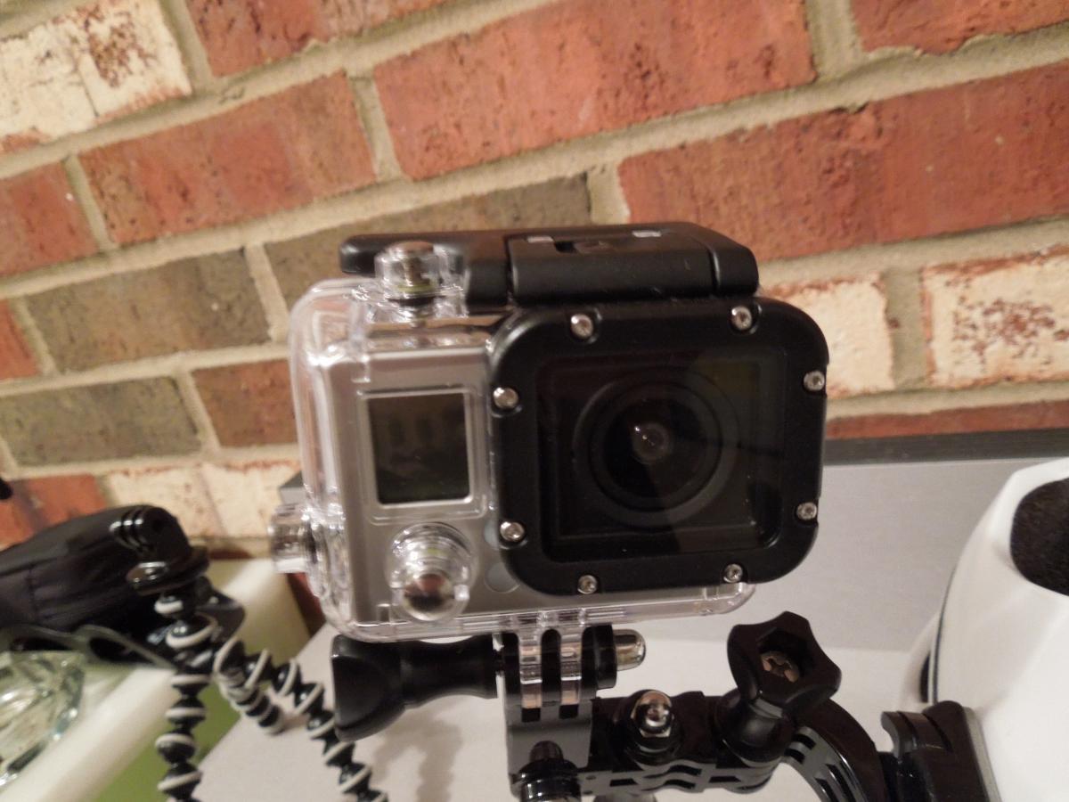 GoPro Hero3 Silver Edition: A close-up of an action camera housed in a clear protective case, mounted on a flexible tripod. The camera features a small LCD screen, various buttons, and a lens visible at the front, with a brick wall in the background.