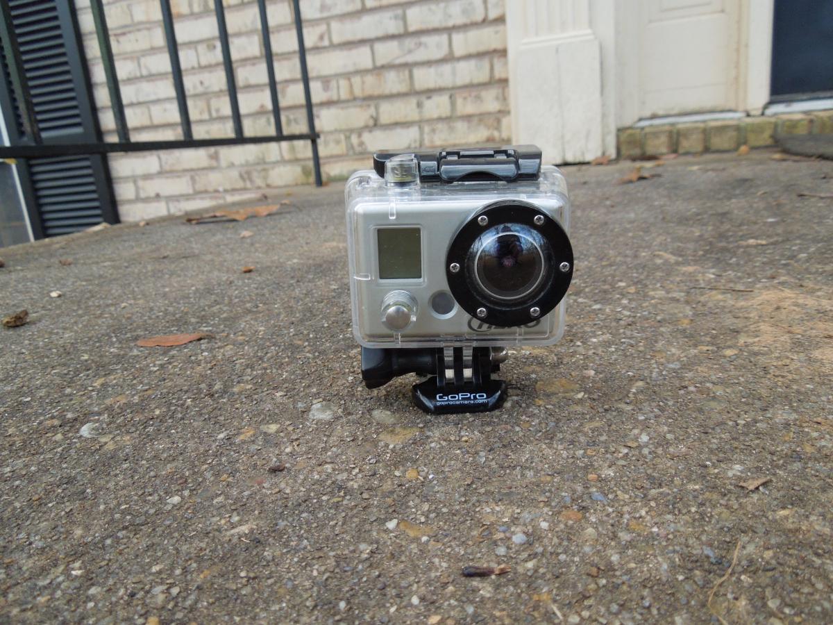 GoPro Helmet HERO: A GoPro camera housed in a clear protective case, positioned on a concrete surface. The camera is facing forward, showcasing its lens and display screen, with a GoPro mount visible underneath. A brick wall and a doorway are blurred in the background.