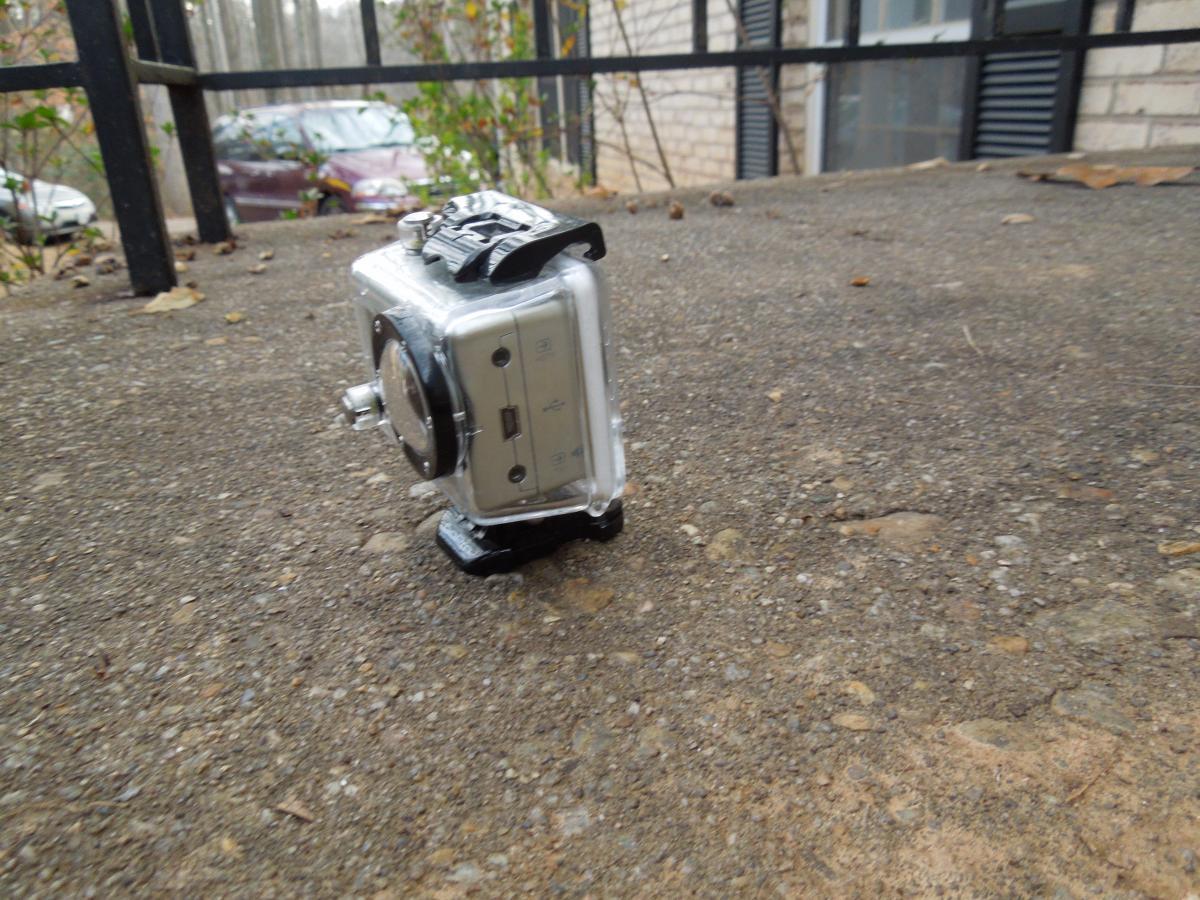GoPro Helmet HERO: A small action camera resting on a concrete surface, partially tilted. The camera has a transparent housing and visible controls on one side. In the background, blurred outlines of parked cars and greenery can be seen, indicating an outdoor setting.