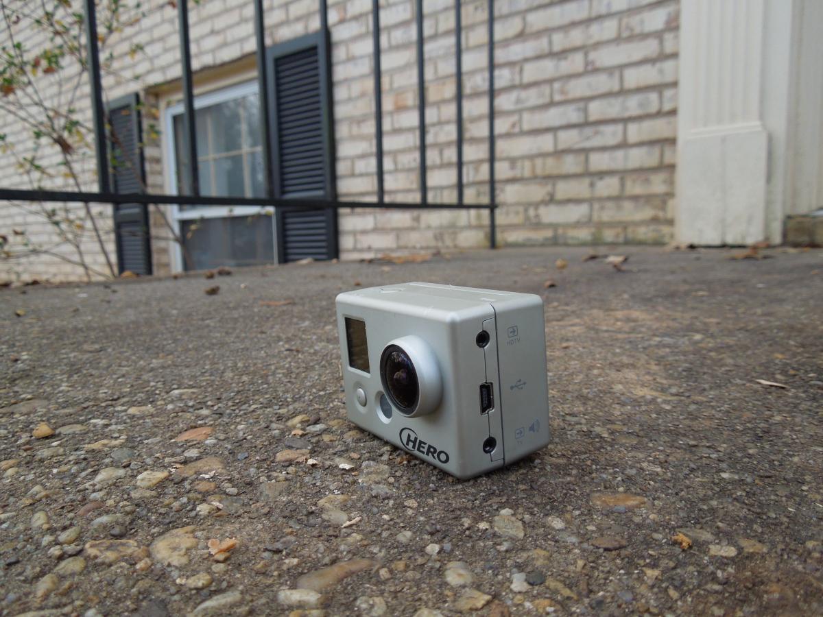 GoPro Helmet HERO: A silver action camera positioned on a textured concrete surface, with the camera lens facing forward. In the background, a window with black shutters can be seen along with a metal railing, hinting at an outdoor setting.