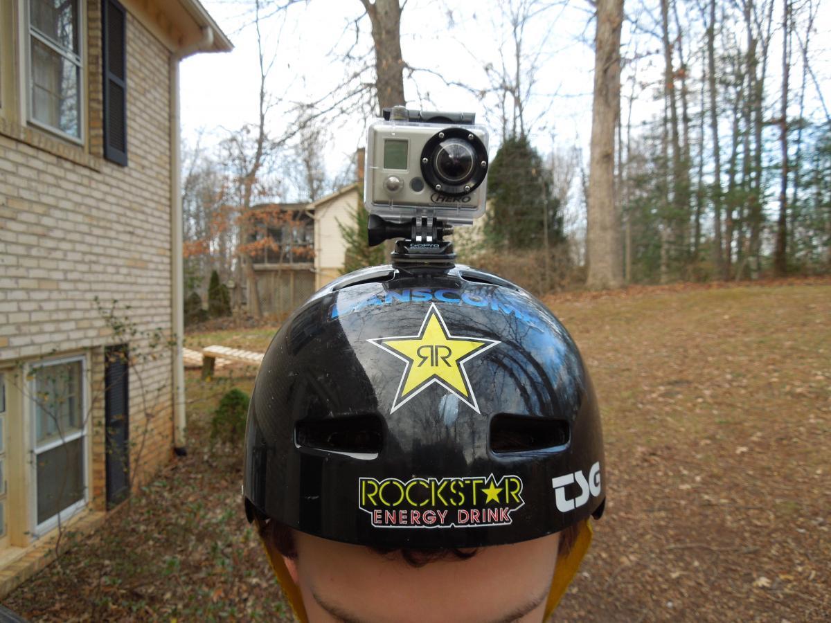 GoPro Helmet HERO: A close-up view of a black helmet adorned with stickers, including a Rockstar Energy Drink logo and a star emblem. A camera is mounted on the top of the helmet, set against a backdrop of trees and a house in a suburban setting.
