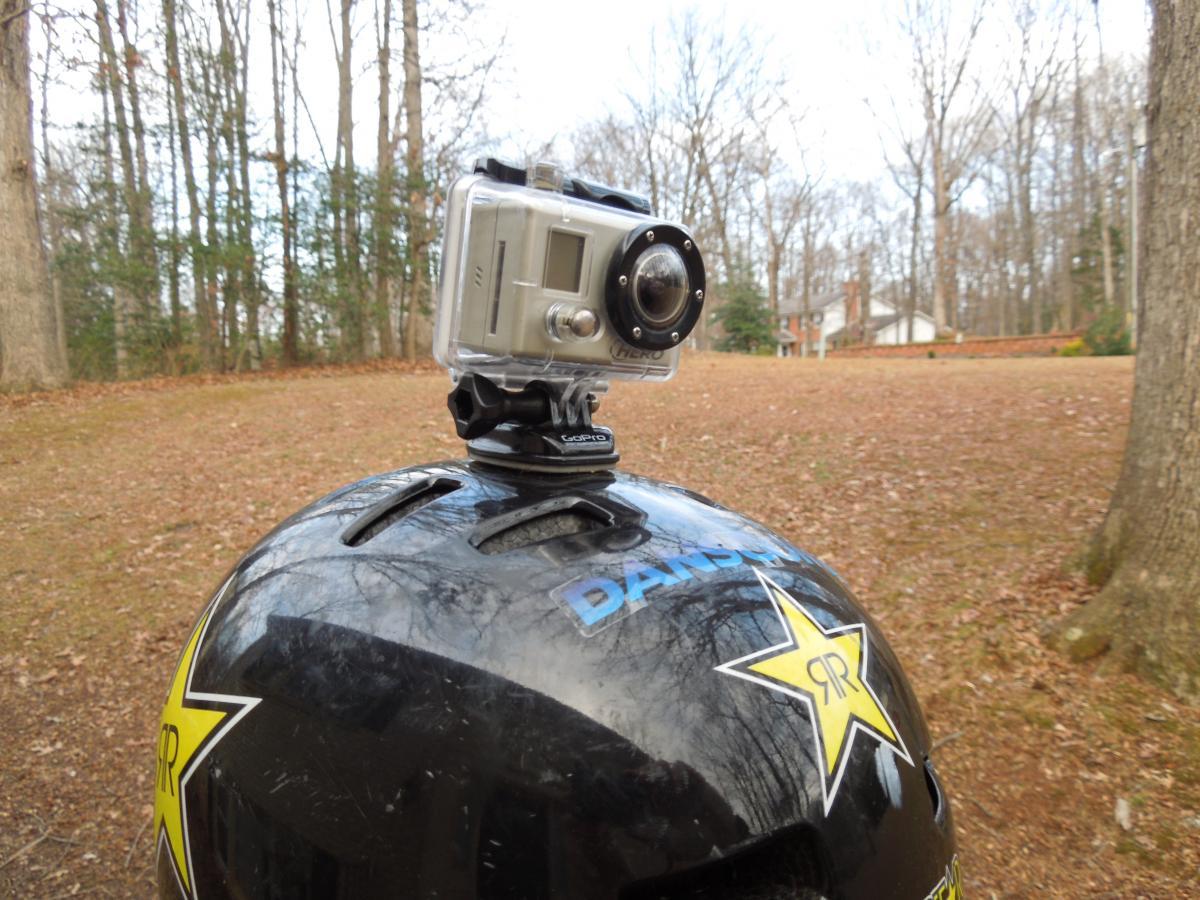 GoPro Helmet HERO: A close-up view of a black helmet with a prominent star logo, featuring a mounted action camera on top. The background shows a wooded area with trees and a grassy ground covered in fallen leaves.