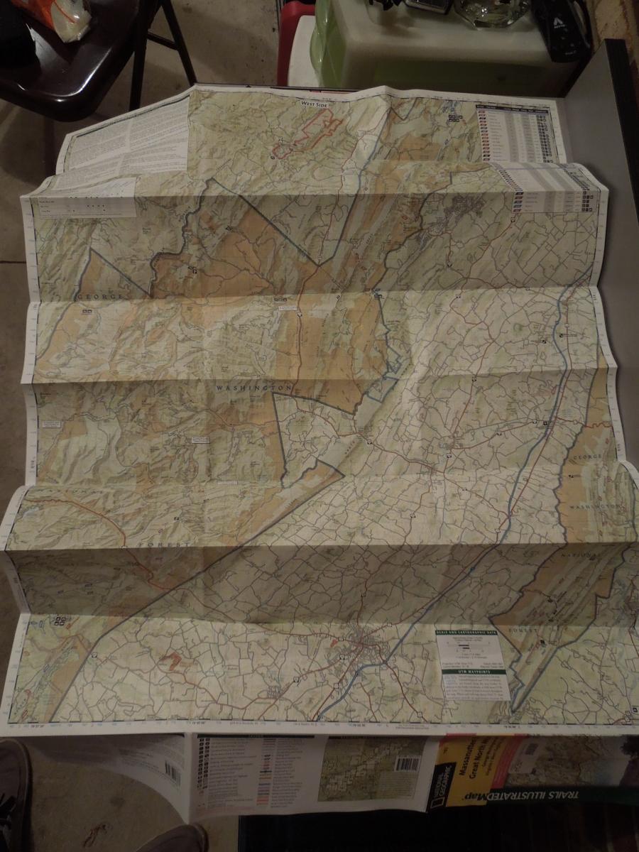 Trails Illustrated National Geographic Massanuten and Great North Mountain: A detailed, unfolded topographic map displaying various geographical features, roads, and landmarks. The map includes shaded relief areas and is oriented with north at the top, with labels indicating states and regions. It is spread out on a flat surface, with a hint of a chair and other items in the background.