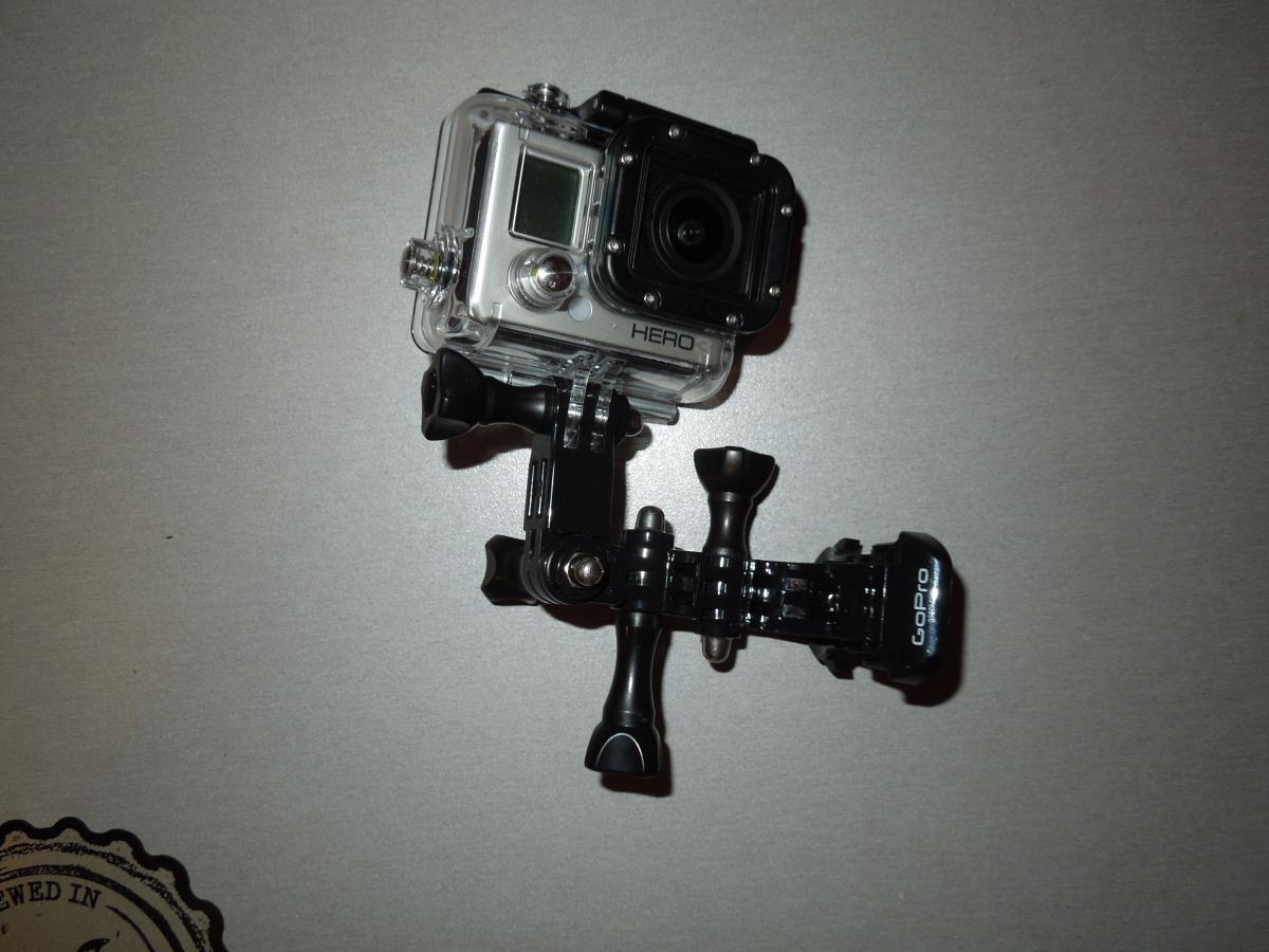 GoPro Helmet Front Mount: A GoPro camera mounted on a black adjustable clamp, positioned on a light gray background. The camera is housed in a clear protective case, displaying the device's screen and lens.