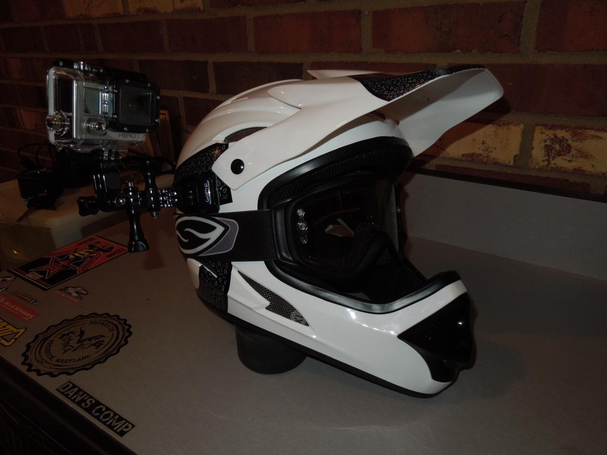 GoPro Helmet Front Mount: A white motocross helmet with a black visor and a camera mounted on the side, positioned on a table with various stickers. In the background, there is a brick wall.