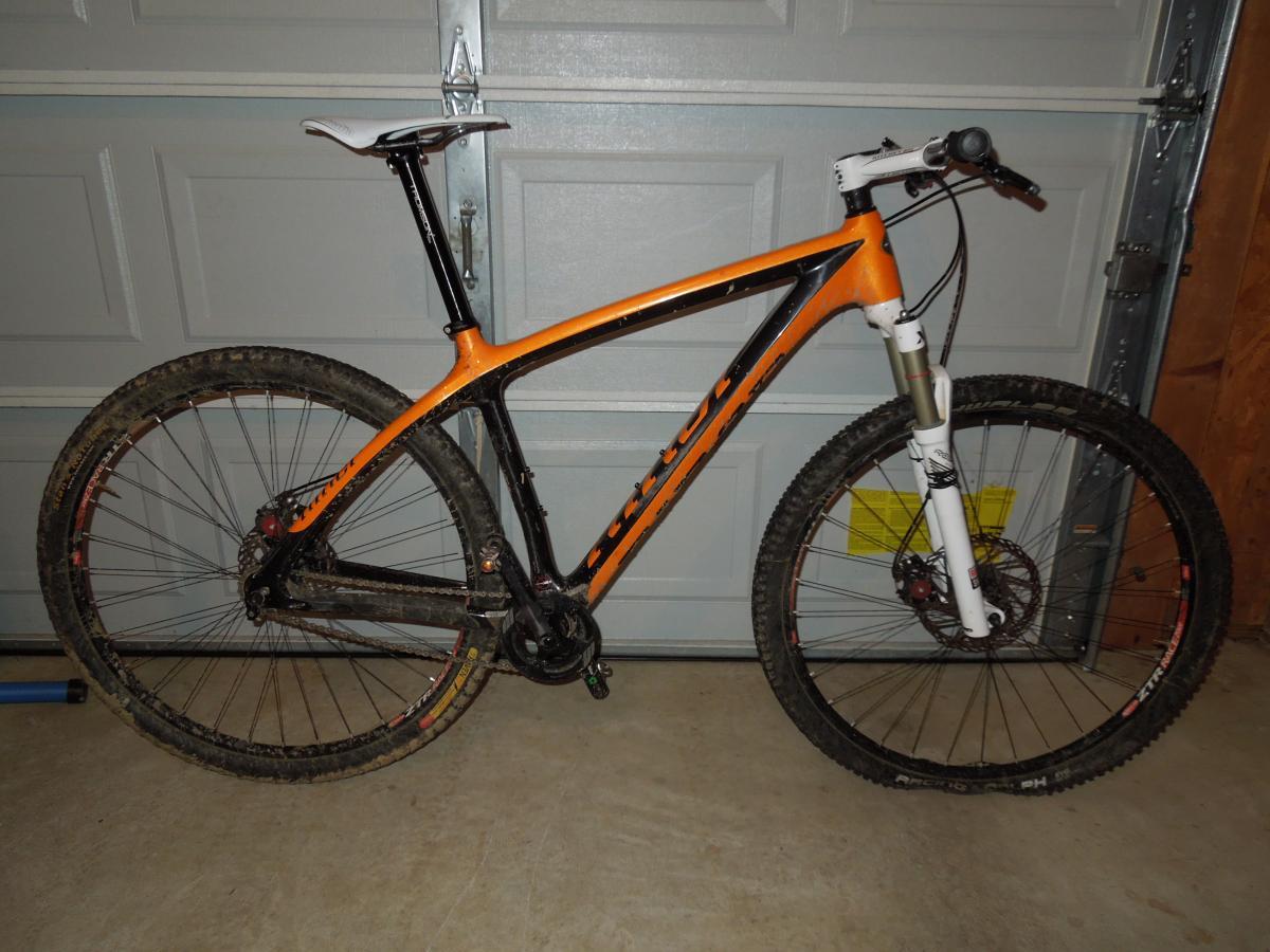 Niner Air 9 Carbon: A mountain bike with an orange and black frame, featuring thick, knobby tires with visible dirt residue. The bike is positioned to the side against a garage door. The front suspension fork is white, and the seat is a sleek design, also primarily white. The bike appears well-used, emphasizing its off-road capabilities.