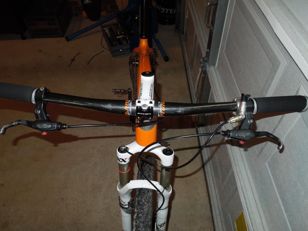 Niner Air 9 Carbon: Top view of a mountain bike's handlebars, featuring a black bar with grips, brake levers, and a visible orange and white frame. The bike is positioned in a garage setting, with concrete flooring and partially visible tools and equipment in the background.