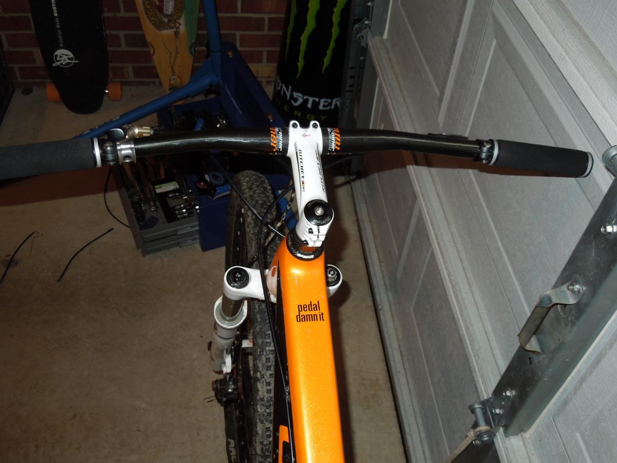 Niner Air 9 Carbon: Alt text: A close-up view of a mountain bike's handlebars and stem, showcasing an orange frame with the words "pedal damnit" printed on it. The background features a garage setting with a skateboard and tools visible.