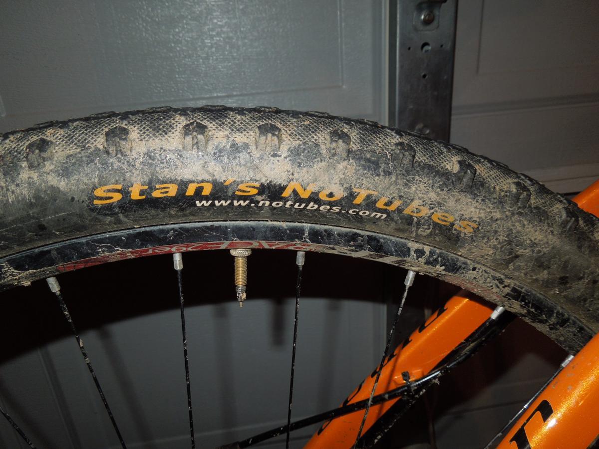 Stans The Raven: Close-up image of a mountain bike tire showing the tread pattern and the label "Stan's No Tubes," with the website "www.notubes.com" visible. The tire is slightly muddy, indicating recent use, and is mounted on an orange bike frame. The background features a garage setting.
