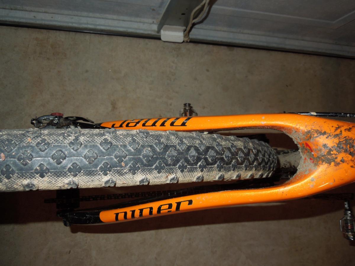Stans The Raven: A top-down view of a mountain bike's front fork and tire, showcasing a worn and muddy tire tread along with the orange branding of the bike's frame. The background is a concrete surface.