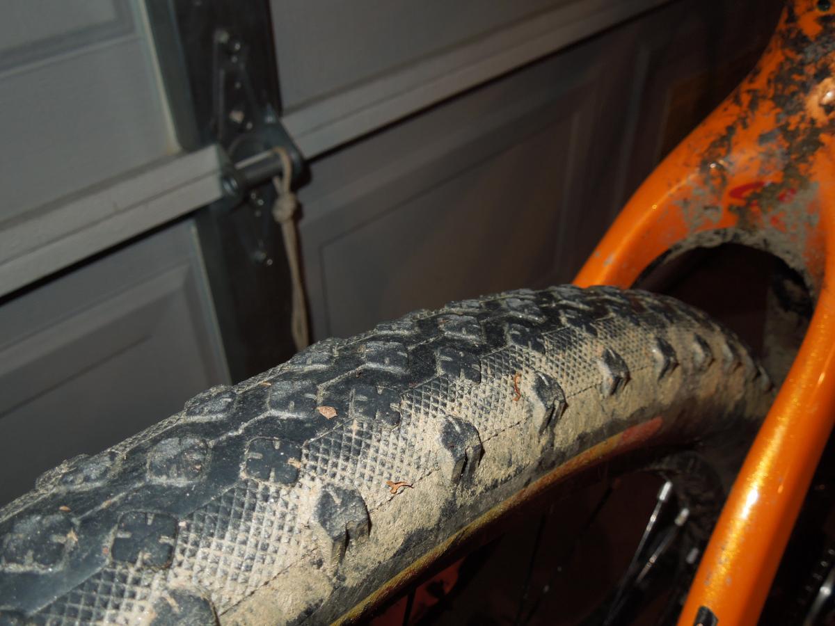 Stans The Raven: Close-up view of a muddy bicycle tire mounted on an orange bike frame, set against a garage background. The tire shows visible tread patterns and dirt accumulation, indicating recent use in off-road conditions.