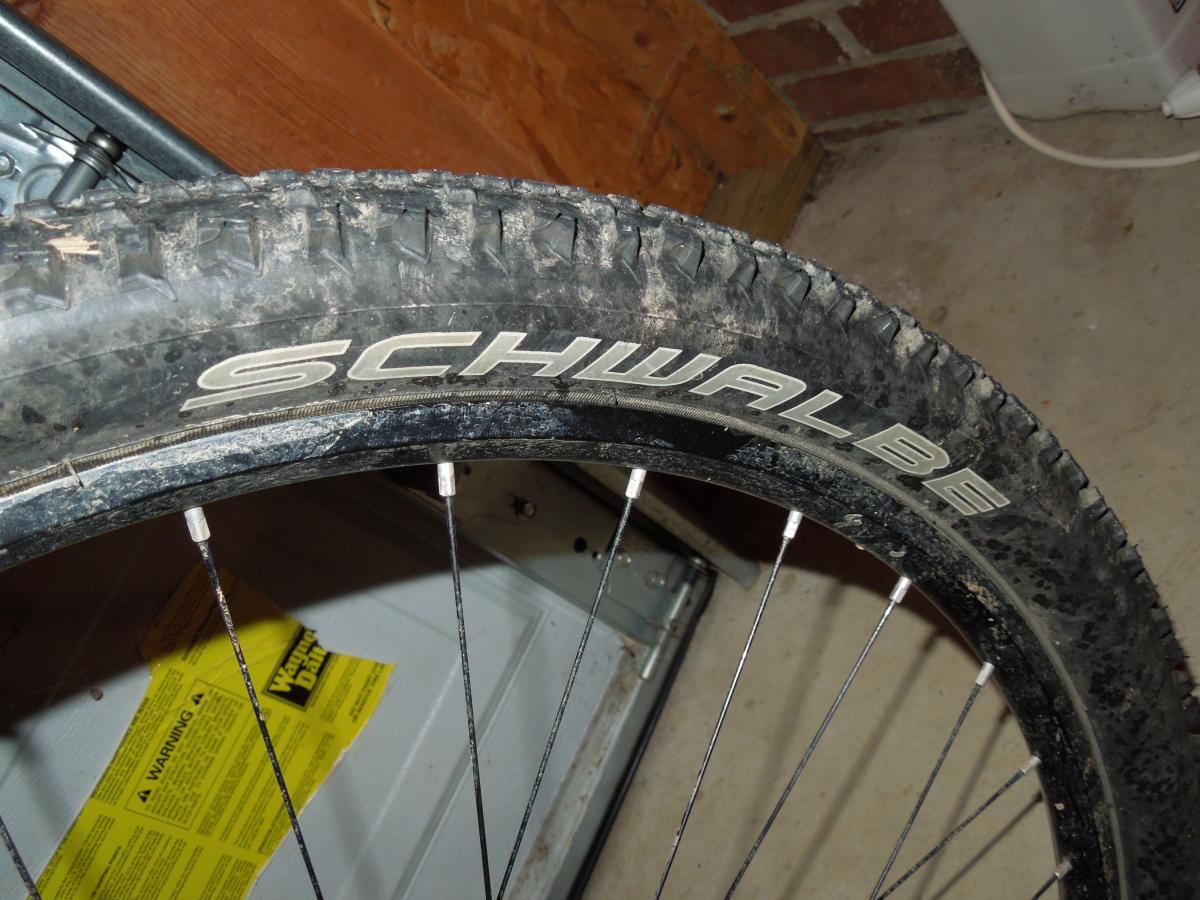 Schwalbe Racing Ralph: Close-up image of a Schwalbe bicycle tire showcasing the tread pattern and logo. The tire appears slightly dirty, indicating it has been used, and is mounted on a wheel with visible spokes. A warning label is faintly visible in the background.