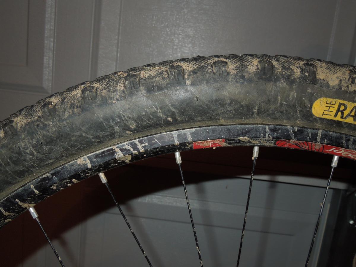 Stans The Raven: Close-up of a dirty mountain bike tire and wheel, showing the tread pattern and a partially worn rim. The image highlights the bike's use, with visible dirt and grime on the rubber.