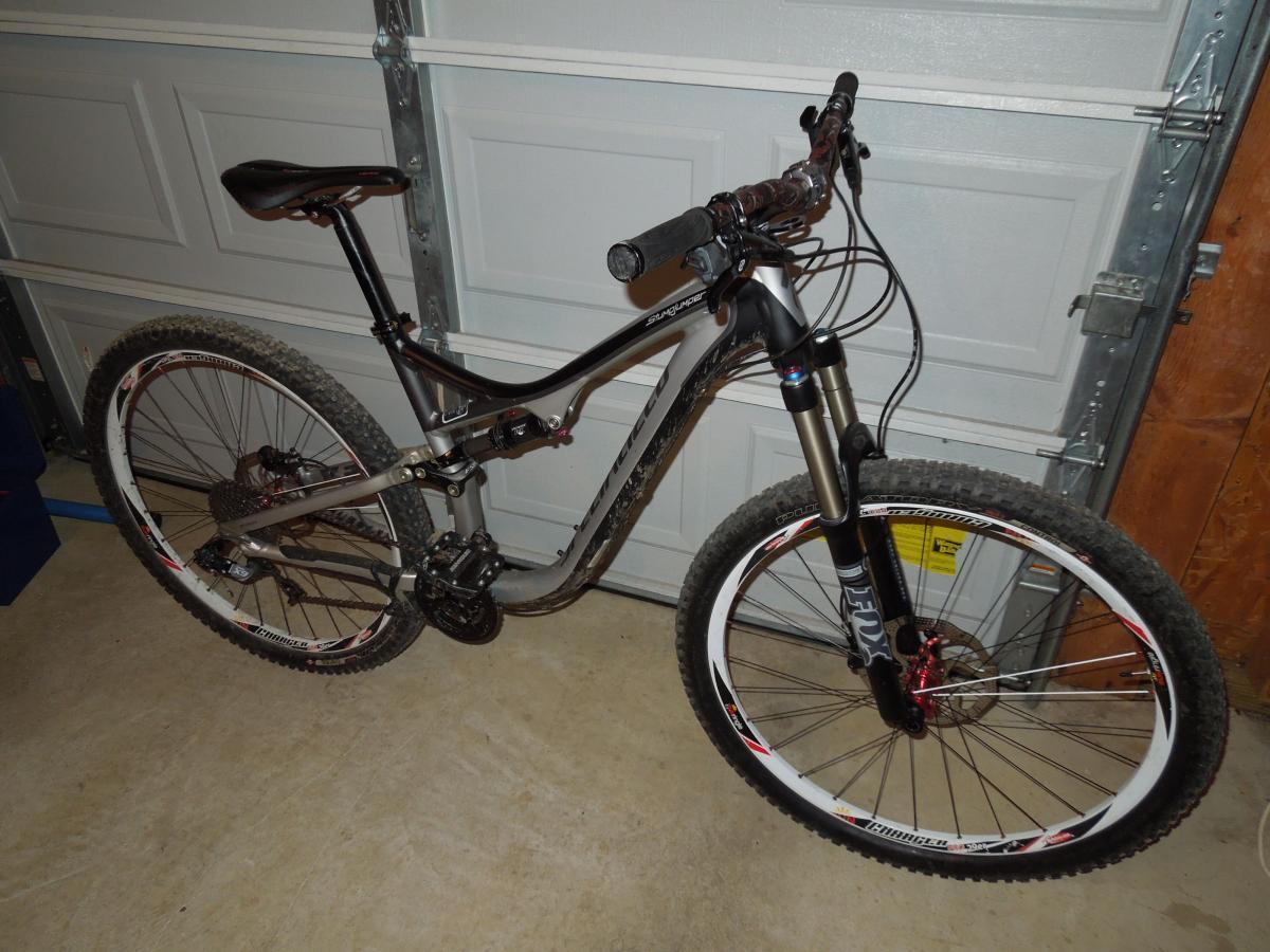 Specialized Purgatory Control 2bliss: Mountain bike resting against a garage wall, featuring a sleek metal frame, knobby tires, and front suspension. The bike has a black and silver color scheme with red accents and is positioned on a concrete floor.