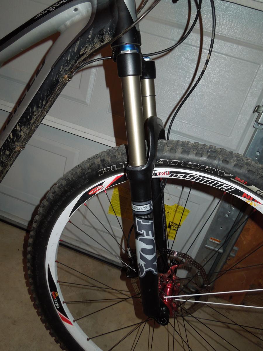 Jagwire Ripcord Brake Kits: Close-up view of a mountain bike's front fork and wheel, showcasing a FOX suspension fork, dirty frame, and tire tread. The background shows a garage setting with a partially visible garage door.