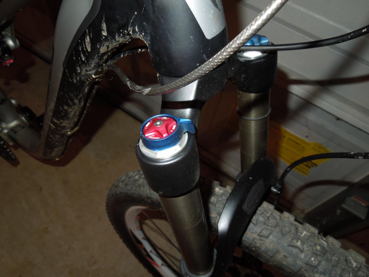 Jagwire Ripcord Brake Kits: Close-up view of a mountain bike fork with suspension adjustment knobs, featuring a blue and red color scheme. The bike shows signs of use, with mud on the frame and tire. The background is a garage setting.