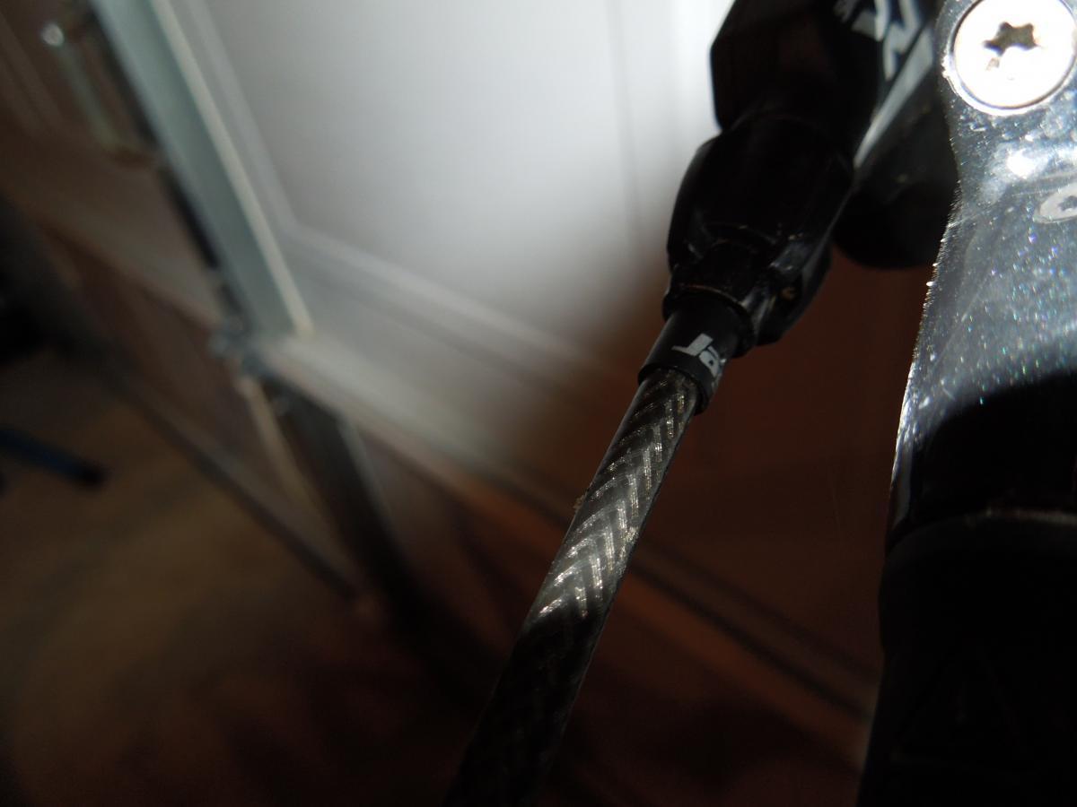 Jagwire Ripcord Brake Kits: Close-up image of a carbon fiber cable with a black connector, partially illuminated, set against a blurred garage background. The focus is on the textured surface of the cable and the details of the connector.