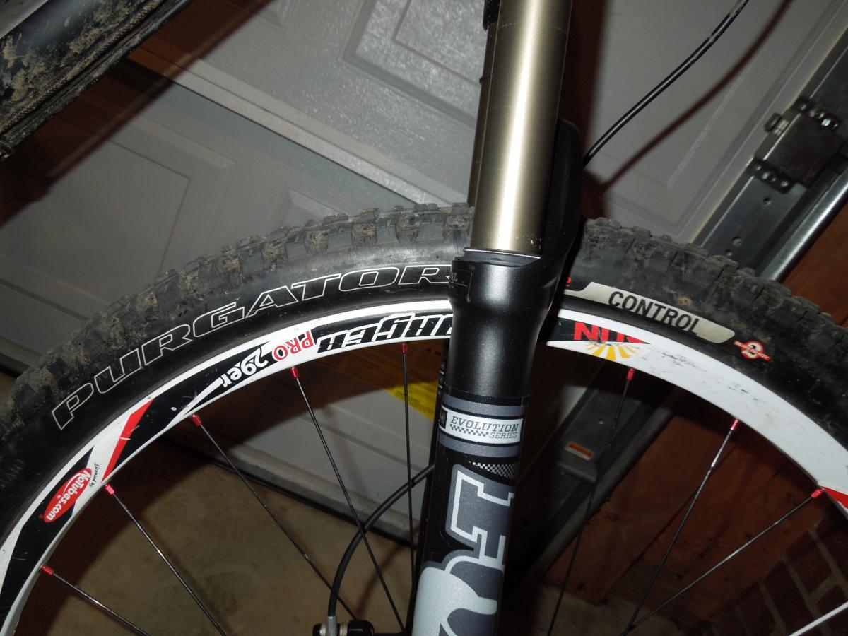 Specialized Purgatory Control 2bliss: Close-up image of a mountain bike front fork and tire. The tire features the "Purgator" model with visible tread and design details. The fork has a marked section labeled "Evolution Series" and showcases a metallic finish, while the background includes a garage setting with tools and equipment.