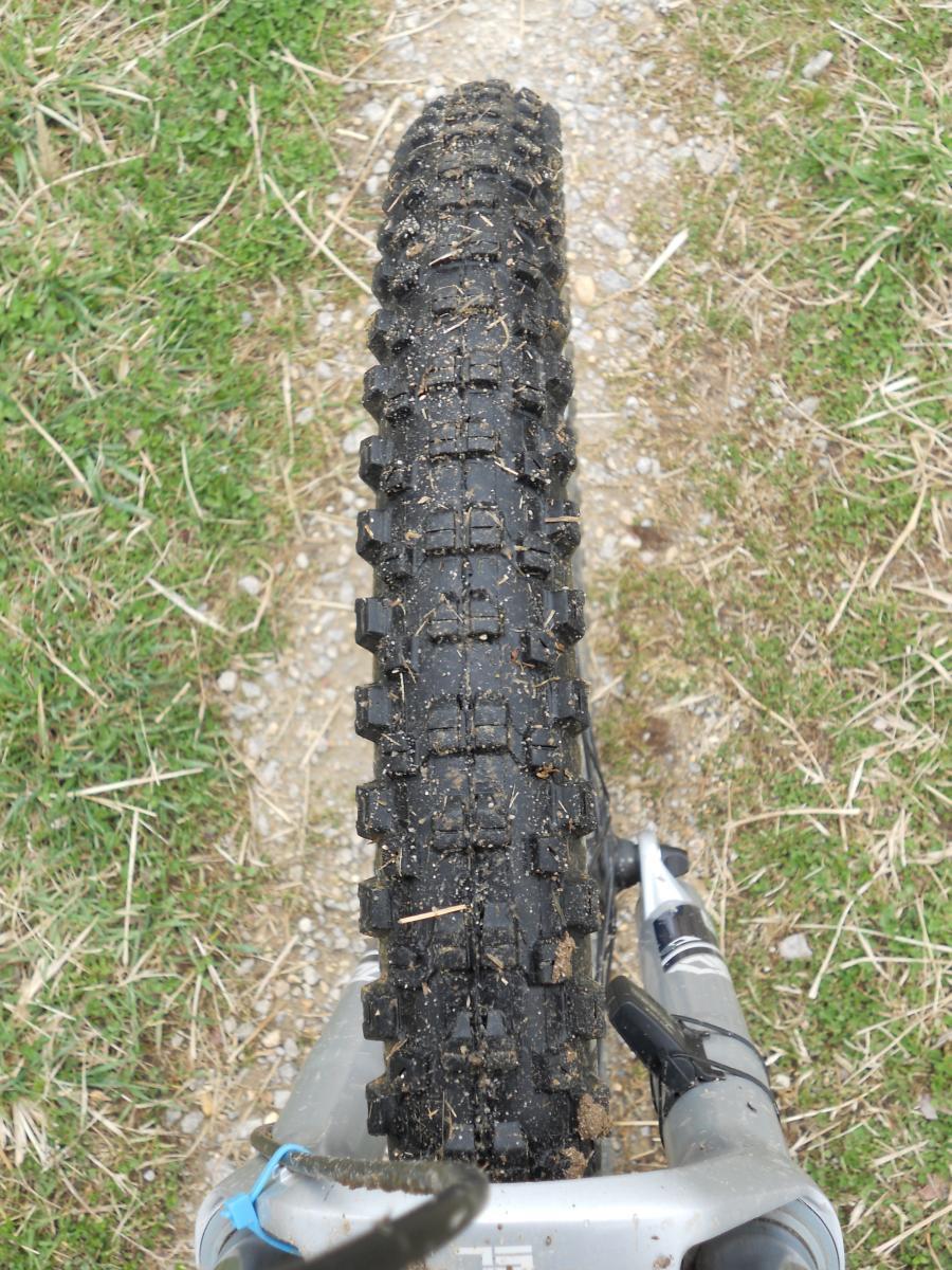 Kenda Nevegal: A close-up view of a mountain bike tire resting on a gravel path, showcasing the tread pattern and some dirt and grass debris on the tire. The bike frame is partially visible, with the image focusing on the tire and surrounding terrain.