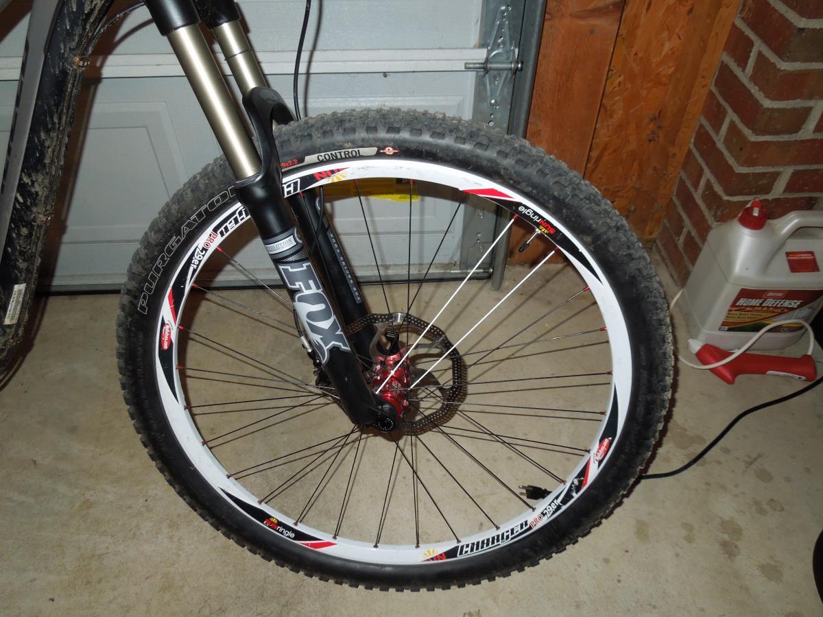 Specialized Purgatory Control 2bliss: Close-up view of a mountain bike's front wheel and suspension fork, showcasing a tire with a textured tread and a white rim. The bike is positioned indoors against a garage door, with some visible floor and background elements, including a container.