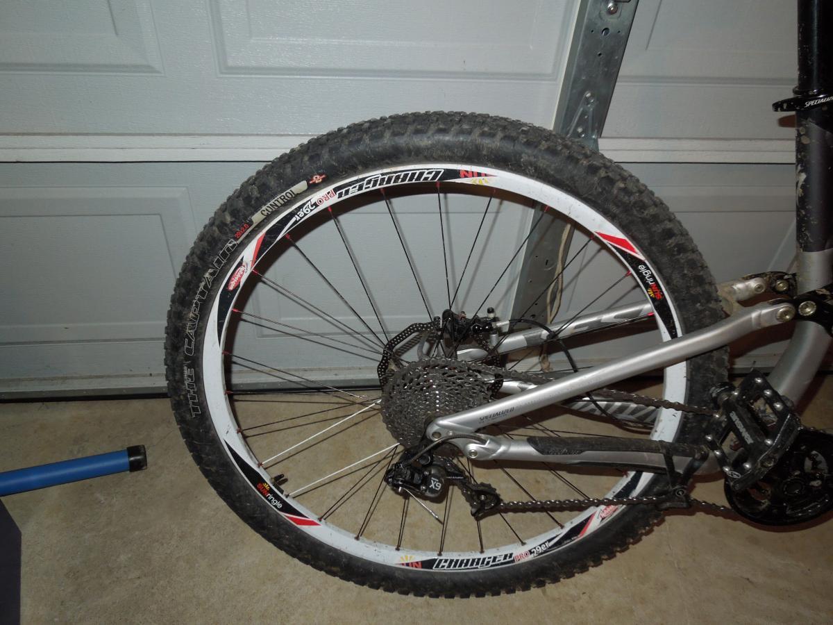Specialized The Captain Control 2bliss: Alt text: Close-up view of a mountain bike's rear wheel and drivetrain, showing the tire tread, cassette, and chain on a garage floor.