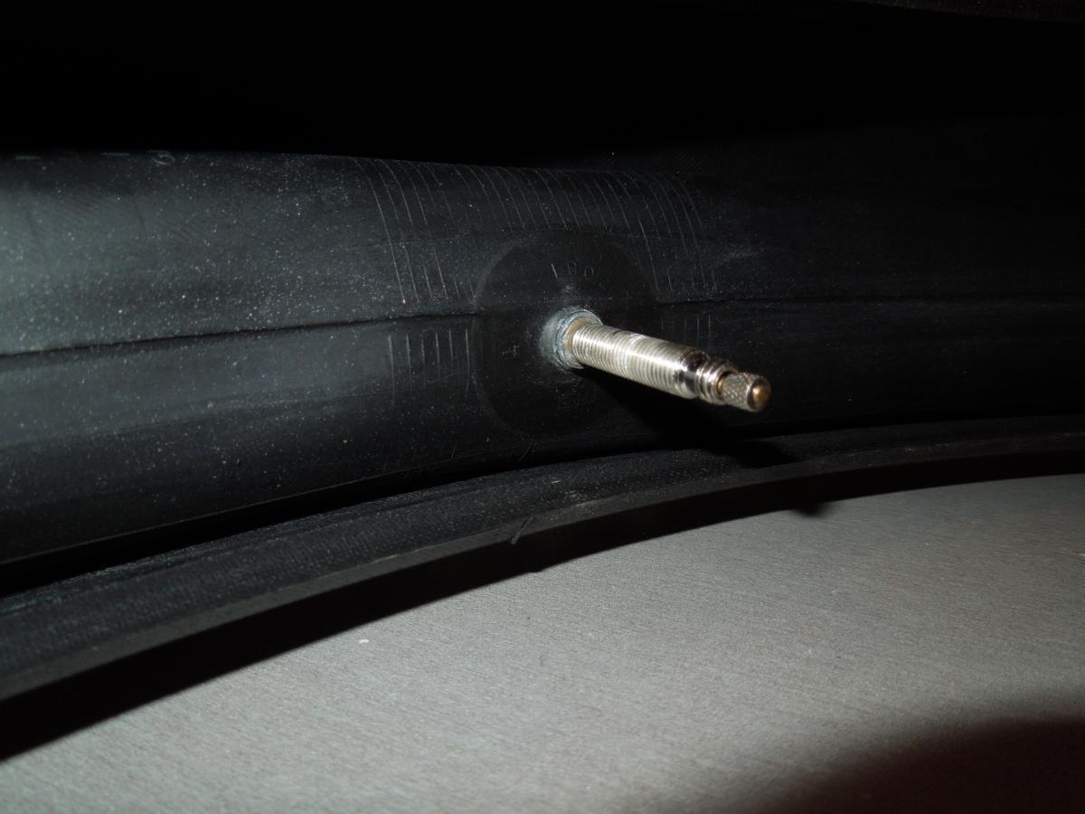 Kenda Downhill Tube: A close-up image of a bicycle tire inner tube showing the valve stem. The tube is black and slightly dusty, resting on a flat surface. The valve stem is metallic with a threaded end, positioned at the center of the image.