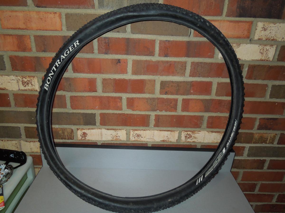 Bontrager 29-3: A black Bontrager bicycle tire mounted on a rim, positioned vertically against a brick wall. The tire features a tread pattern suitable for off-road use, with visible brand markings. The background includes a light-colored surface and a glimpse of other objects.