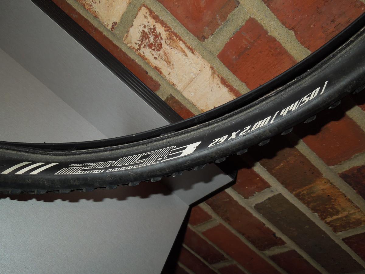 Bontrager 29-3: A close-up image of a bicycle tire mounted on a wall, displaying tire specifications including "29 x 2.00" and "44/50" in white text against a black background. In the background, there are brick and gray surfaces.